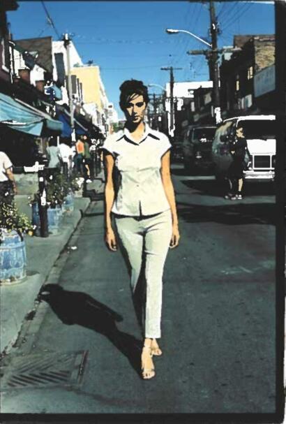 A woman in white walking on a city street under utility lines, with shops and parked cars in the background.
